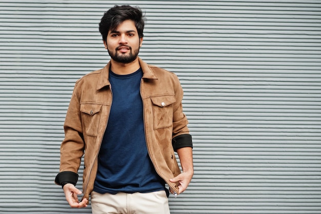 Stylish casual indian man wear blue tshirt and brown jacket posing against grey wall