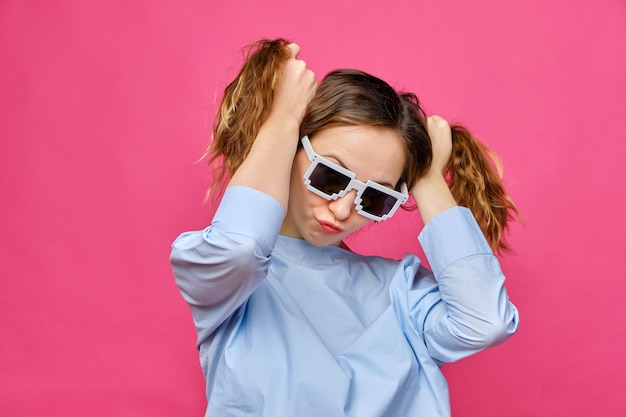 Stylish caucasian girl in a pale blue t-shirt and 8-bit glasses and makes two tails of hair on a pink background.