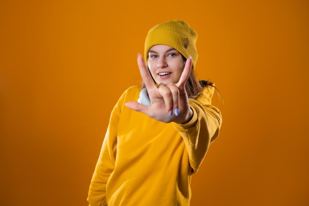 Stylish cheerful young brunette in a yellow hoodie and hats shows a cool gesture two fingers up on a...