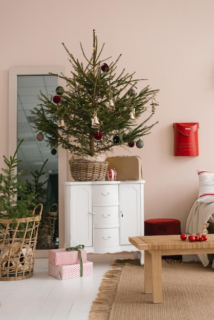 Stylish Christmas tree with toys in a wicker basket stands on a white chest of drawers in interior