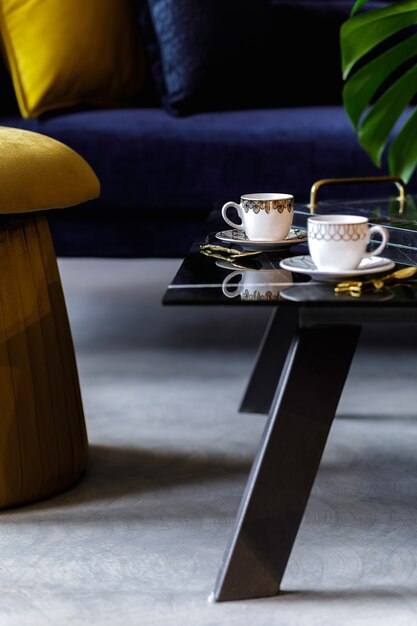 Stylish composition at living room interior with gold pouf, velvet sofa, pillows, coffee table, tropical leaf and cups of coffee in modern concept