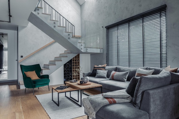 Stylish composition of modern living room design with corner grey sofa green armchair coffee table and minimalist personal accessories Stairs in living room interior Concrete walls Template