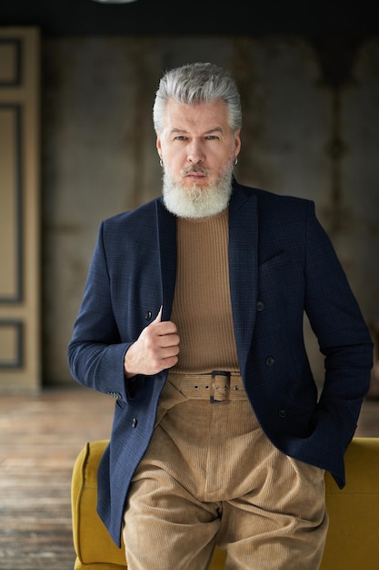 Stylish confident mature man in casual clothes looking at camera while posing leaning on sofa indoors