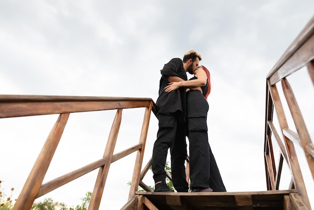 Stylish couple in love in black clothes walks in the park and hugs A lovely couple of hipsters are standing on a wooden bridge and kissing Youth love and lifestyle concept