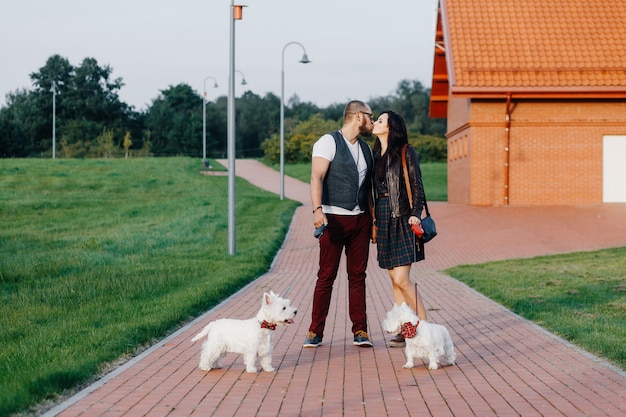 A stylish couple strolls through the park with two white dogs