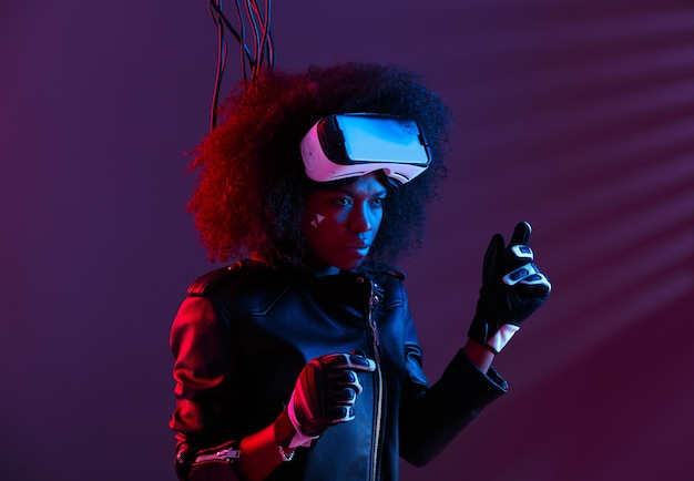 Stylish curly dark haired girl dressed in black leather jacket and gloves is wearing the virtual reality glasses on her head in the dark studio with neon light . Cyberpunk character