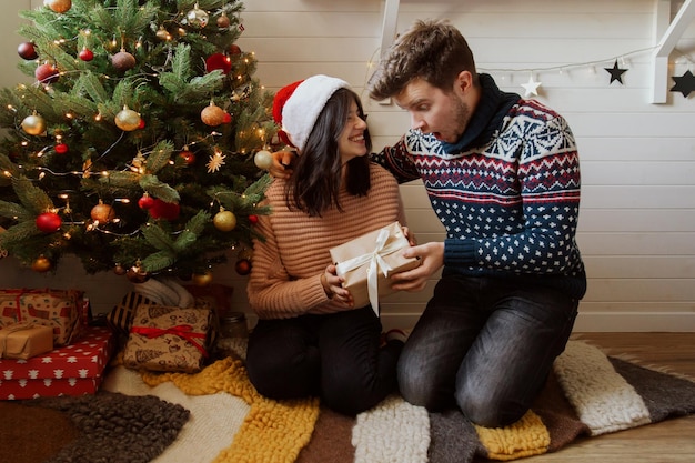 Stylish emotional couple exchanging christmas gifts under christmas tree in festive room