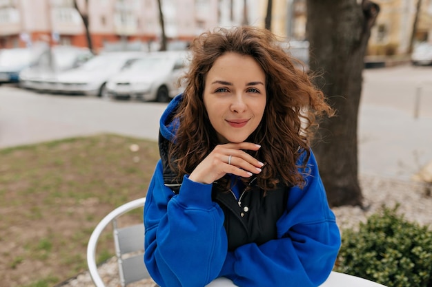 Stylish european woman with curly hairstyle wearing blue pullover posing at camera while sitting on summer terrace in city Lifestyle portrait of young caucasian girl laughing outdoors