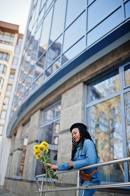 Stylish fashionable african american women in jeans wear and black beret with yellow flowers bouquet posed outdoor in sunny day against blue modern building