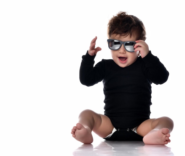 Stylish fashionable boy in black overalls sunglasses sitting on the floor Smiling positive toddler child Isolated portrait shot against a white studio background Happy childhood style and fashion