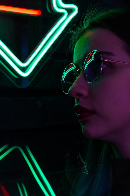 Stylish fashionable teenage girl in glasses with reflection looks up on the street with neon illumination of the city Bar and music concept