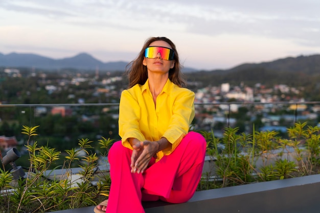 Stylish fit fashion women in bright pink wide leg pants and yellow shirt holding bag trendy sunglasses posing at rooftop terrace tropical view outdoor natural day light