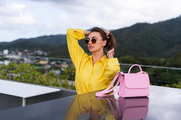 Stylish fit fashion women in bright yellow shirt trendy sunglasses posing at rooftop terrace tropical view outdoor with pink bag