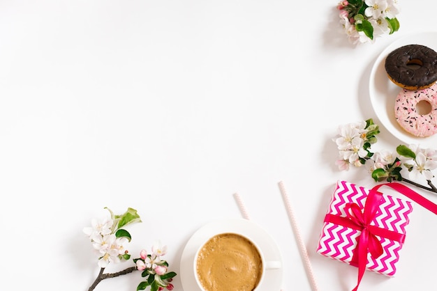 Stylish flat lay with a cup of coffee apple blossoms donuts and a gift box of birthday or valentine's sweets on a white background with copy space