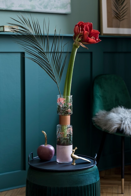 Stylish and floral composition of beautiful flowers in modern vase on the velvet pouf with elegant accessories and furnitures. Blossom concept at living room. Green walls. Interior design. Template.