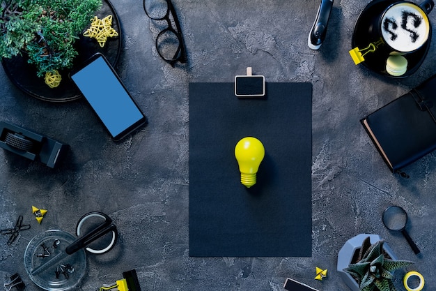 Stylish freelancer workspace. Home office desk table flat lay. Yellow idea lightbulb on paper page, smartphone blank screen and stationary top view.