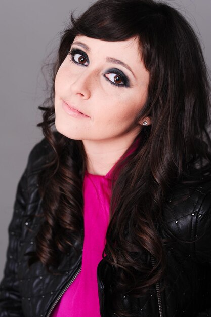 Stylish girl 20-25 year old wearing trendy leather jacket and pink dress posing in studio over gray.