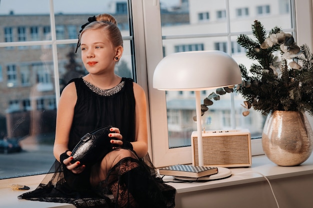 Photo a stylish girl in a black dress sits on the windowsill by the window with a purse in her hands.