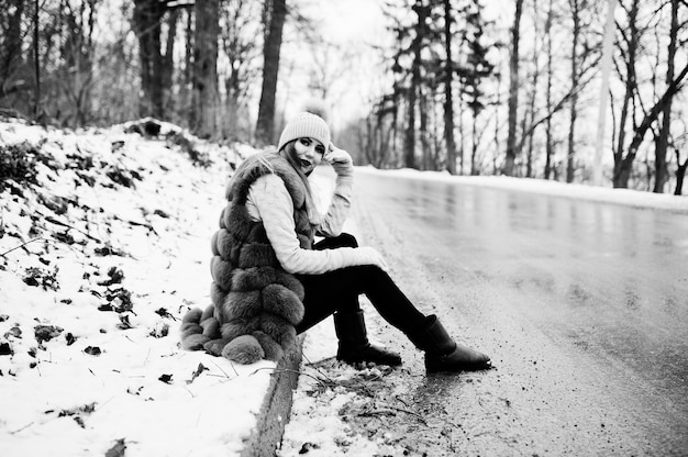 Stylish girl in fur coat and headwear at winter day on sitting border ofroad.