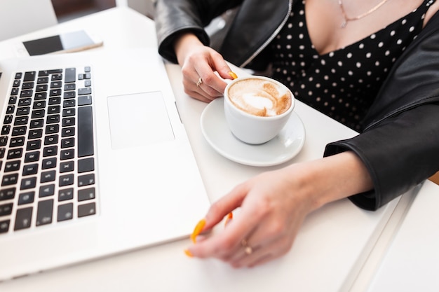 Stylish girl in glamorous fashionable black outfit works at computer sitting at white table in cafe. Successful woman typing on laptop and drinking coffee. Close-up of female hands. Top view.