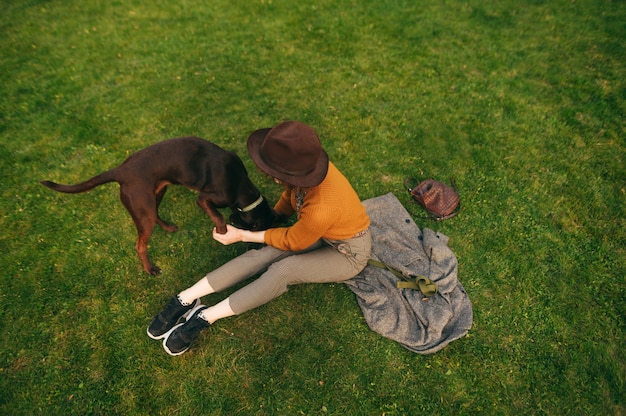 Stylish girl in a hat sits on a green lawn and plays with a brown dog