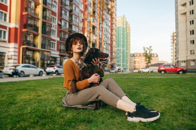 Stylish girl in a hat and vintage clothes sits on the green grass in the yard and hugs the dog