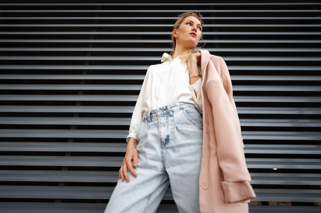 Stylish girl in jeans and white cotton blouse posing near gray wall