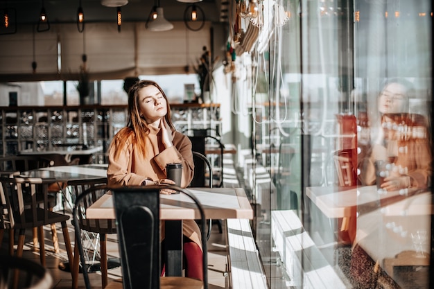 Stylish girl sits in a cafe at the table and drinks coffee. Coffee to go in a cardboard cup. Woman with red hair in a beige warm suit in a cozy atmosphere. Modern interior. Calm and pleasant pastime.