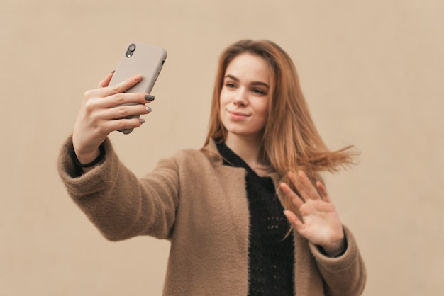 Stylish girl in warm clothes takes selfie spring on the background of beige wall