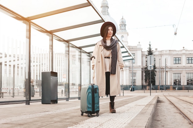 Stylish girl with a suitcase stands at a city stop awaiting a tram