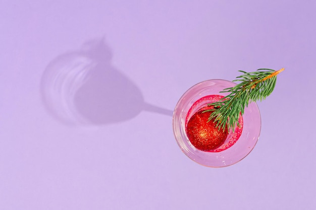 Stylish glass with a pink refreshing drink. Pink tones and reflections. Christmas cherry decoration made of pine twigs as a decoration in a glass. Pastel purple background. Flat lay with soft shadow.