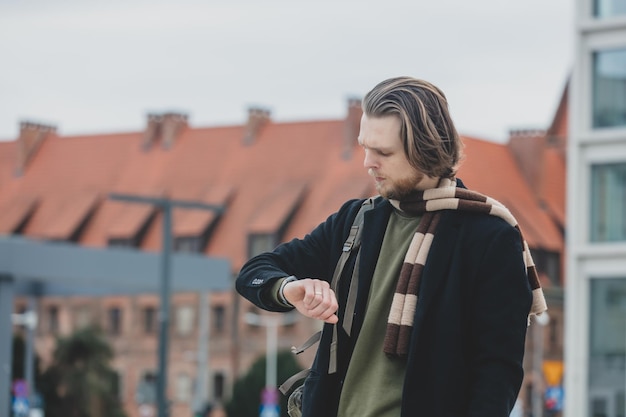 Stylish guy in scarf and coat checks the time on his watch standing at street of Wroclaw Poland