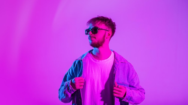 Stylish handsome hipster fashion man with sunglasses in denim fashion clothes with jacket and tshirt posing in studio with creative colorful pink and blue light Trendy portrait