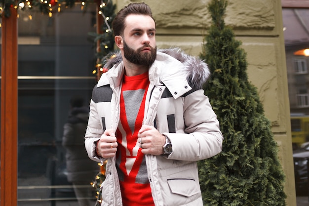 Stylish handsome man with a beard in a winter jacket with fur posing outdoors.