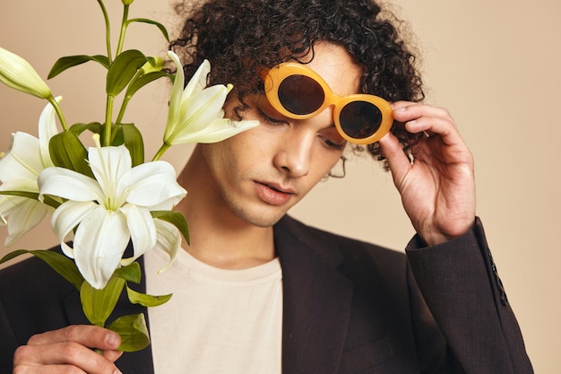 Stylish handsome tanned curly man in trendy sunglasses hold lilies posing isolated on over beige pastel background Fashion New Collection offer Retro style concept Free place for ad