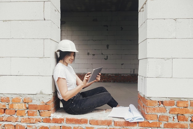 Stylish happy woman engineer in hard hat looking at digital plans on tablet while sitting in window of new modern house Young female architect checking blueprints at construction site