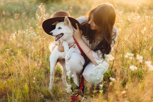 Stylish happy woman playing with cute dog with hat among wildflowers in sunset light Summer travel with pet Young carefree female having fun with white danish spitz in summer meadow