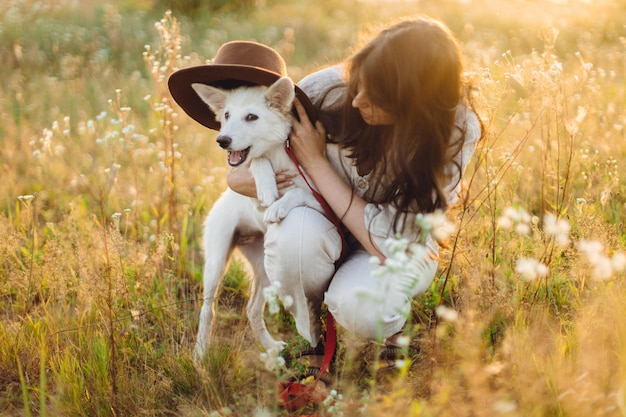 Stylish happy woman playing with cute dog with hat among wildflowers in sunset light Summer vacation with pet Young carefree female relaxing with white danish spitz in summer meadow