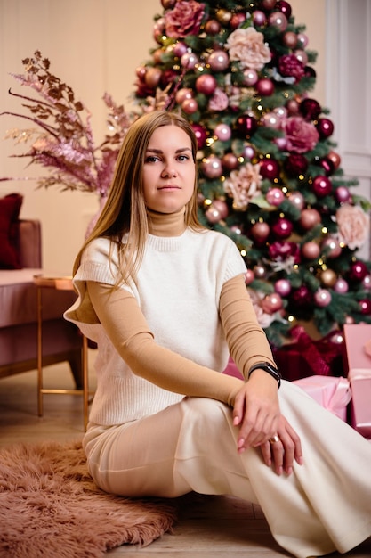 A stylish happy young woman in a fashionable white knitted suit is sitting on a soft rug in a cozy room near a Christmas tree Fashionable girl joyful model rests in the Christmas studio