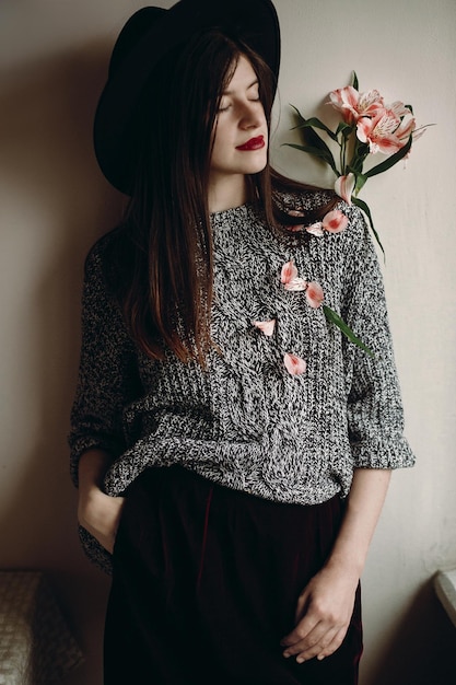 Stylish hipster girl in hat holding pink flowers in room boho woman holding beautiful alstroemeria in hands in spring morning and petals on body creative sensual female portrait