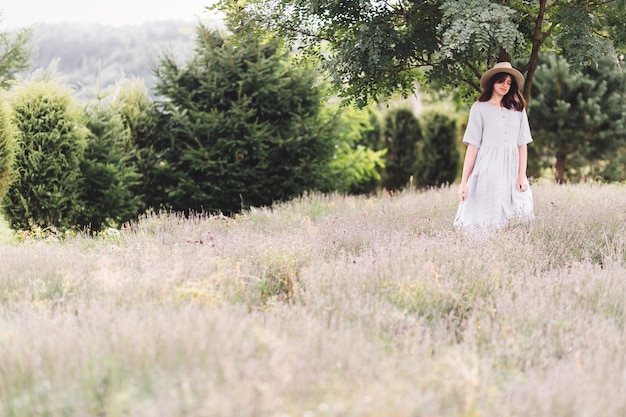 Photo stylish hipster girl in linen dress and hat relaxing in lavender field near tree focus on lavender bohemian woman enjoying summer in mountains atmospheric calm rural moment space for text