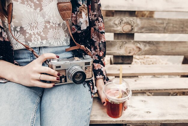 Photo stylish hipster woman holding old photo camera and lemonade boho girl in denim and bohemian clothes sitting on wooden bench at street food festival summer travel space for text