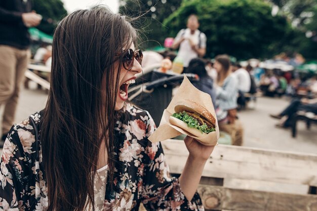 Photo stylish hipster woman in sunglasses eating juicy burger boho girl holding cheeseburger and smiling at street food festival summertime summer vacation travel space for text