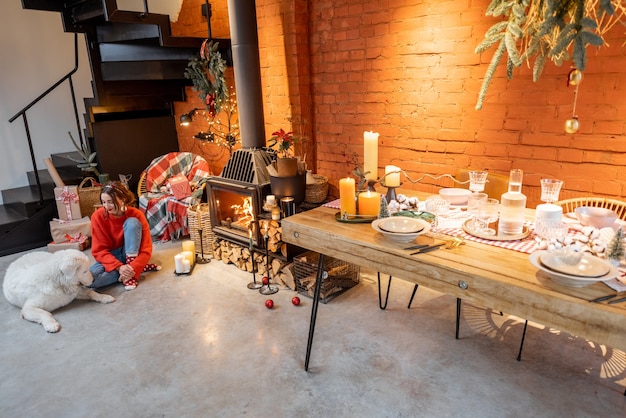 Stylish home interior with festively decorated dining table and fireplace. Woman with a dog during New Year holidays