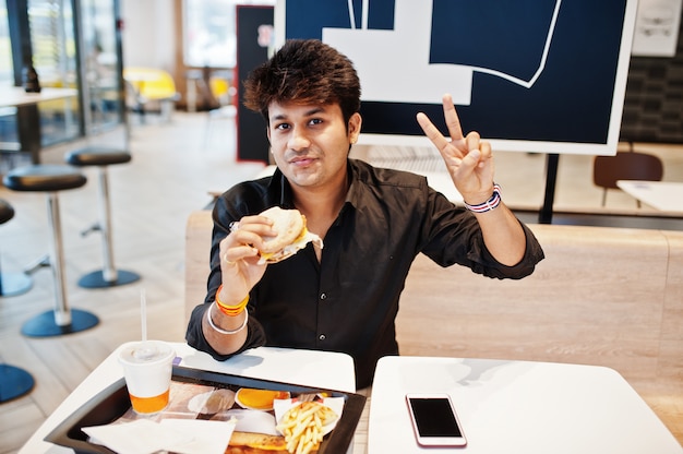 Stylish indian man sitting at fast food cafe and eating hamburger and gesture peace sign hand.