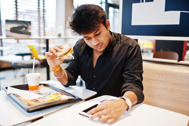 Stylish indian man sitting at fast food cafe and eating hamburger read morning news on mobile phone