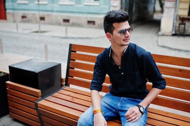 Stylish indian model man in casual clothes black shirt and sunglasses posed outdoor at street of India and sitting on bench