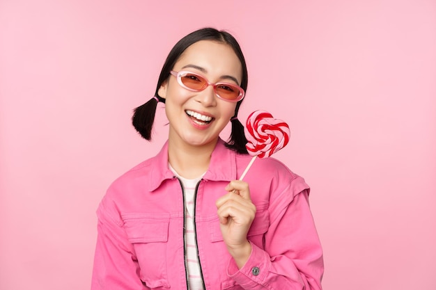 Stylish korean girl licking lolipop eating candy and smiling standing in sunglasses against pink background