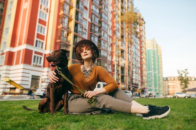 Stylish lady in trendy vintage clothes sits on the lawn in the yard with a beautiful dog hugs a pet