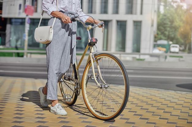 Stylish lady in white standing on the asphalt in the city street in the sunny day while holding a handlebar of a city bicycle